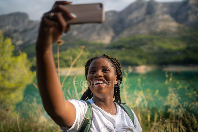 Delighted African American female traveler taking self shot on smartphone on background of lake in highlands in summer — Stock Photo