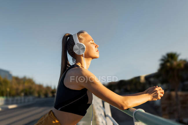 Joven atlético caucásico apoyado en barandilla al aire libre, escuchando música y relajándose con los ojos cerrados después de hacer ejercicio - foto de stock