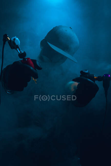 Anonyme stilvolle männliche Tätowierer mit Tätowiermaschinen stehen in dunklen Studio mit Rauch — Stockfoto