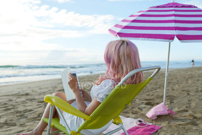 Visão traseira de freelancer feminino anônimo sentado na espreguiçadeira e trabalhando em tablet na praia de areia perto do mar no verão — Fotografia de Stock