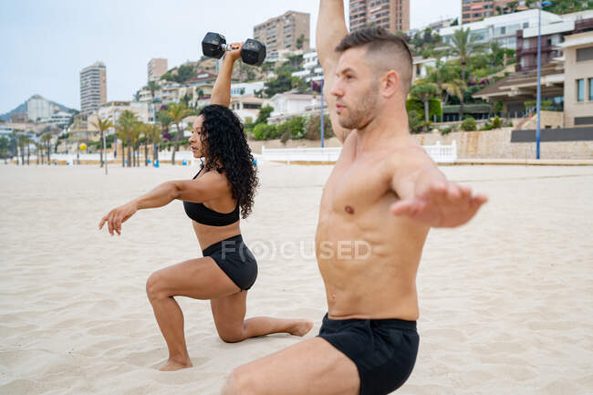 Vista lateral de deportistas multiétnicos musculosos que hacen ejercicio con pesas mientras entrenan en la playa de arena en verano - foto de stock