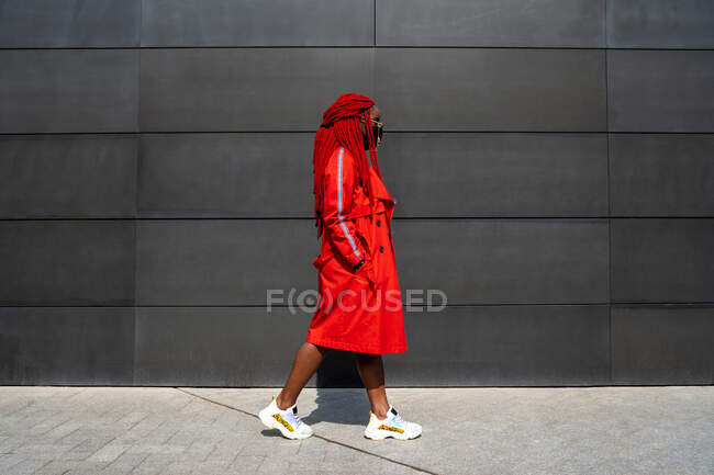 Corps complet de femme afro-américaine en lunettes de soleil marchant avec les mains dans des poches de manteau contre le mur gris — Photo de stock