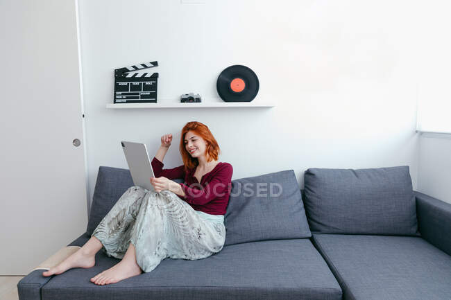 Content young female sitting on sofa while talking to partner during video chat on tablet in house — Stock Photo