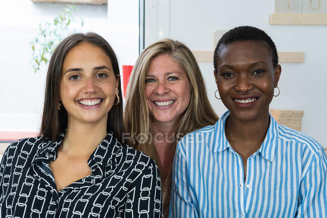 Délicieuses collègues féminines multiraciales regardant la caméra et souriant tout en étant assis sur le lieu de travail ensemble — Photo de stock