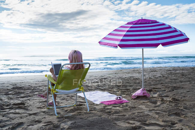 Vista trasera de una freelancer anónima sentada en una tumbona y trabajando en una tableta en la playa de arena cerca del mar en verano - foto de stock