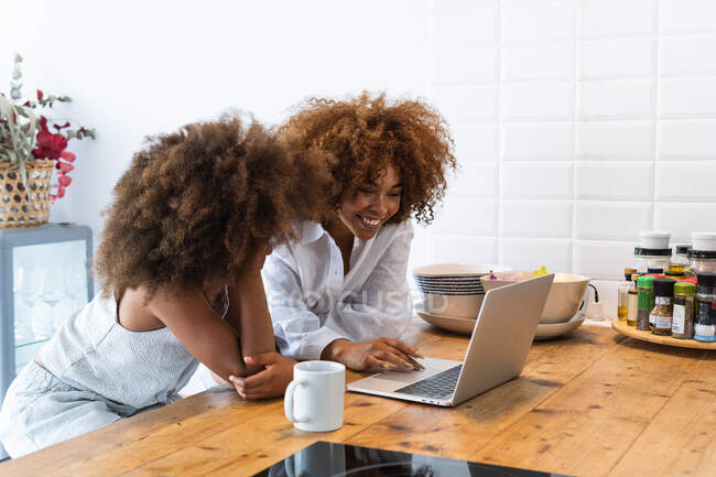 African American woman and girl with Afro hairstyle sitting at kitchen table and browsing netbook together in weekend morning at home — Stock Photo