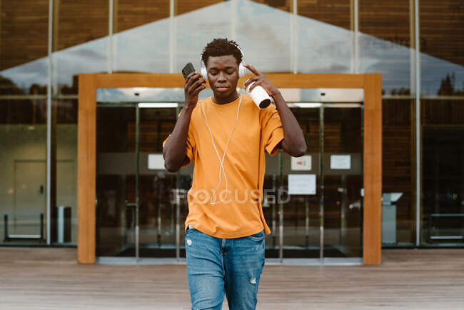 Serious African American male with smartphone listening to music in headphones while walking against modern building — Stock Photo