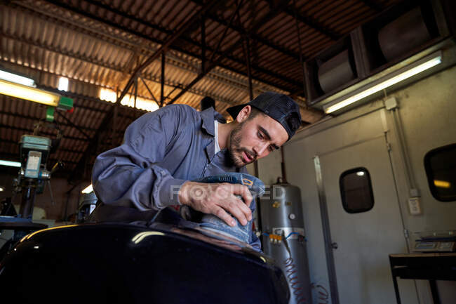 З-під низького кута концентрований чоловік-робітник стоїть на робочому місці і полірує мотоциклетний танк з електричним інструментом під час роботи в гаражі. — стокове фото