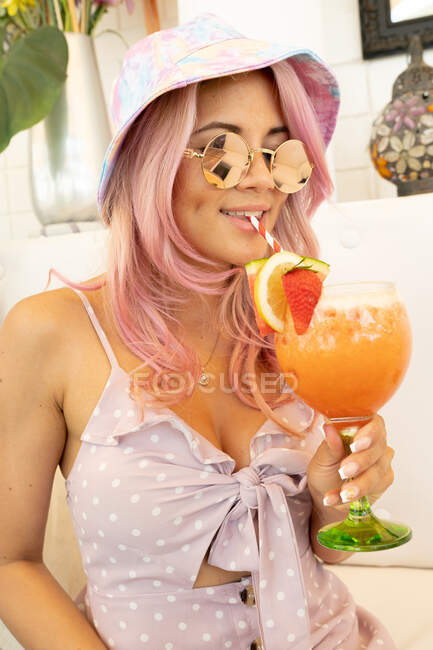 Contenida hembra con pelo rosa y en traje de verano bebiendo cóctel naranja dulce con frutas durante las vacaciones - foto de stock