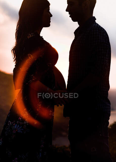 Siluetas de vista lateral de pareja esperando al bebé de pie mirándose cogidos de la mano mientras admiran el atardecer en las montañas juntos - foto de stock