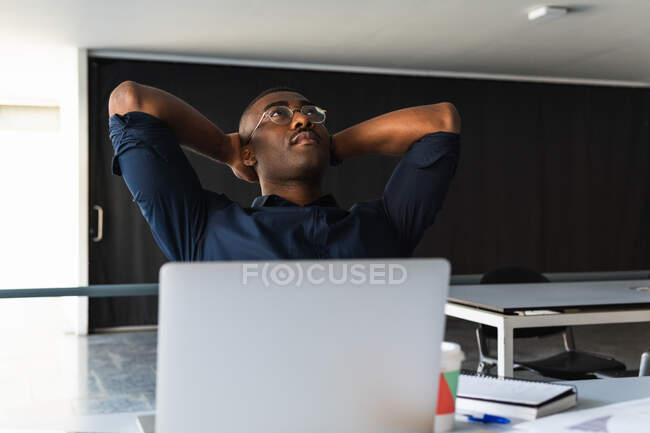 Hombre adulto afroamericano reflexivo en anteojos y ropa formal sentado en la mesa mientras toma un descanso del trabajo cerca de netbook y documentos con bebida para llevar y portátil en el espacio de trabajo ligero - foto de stock