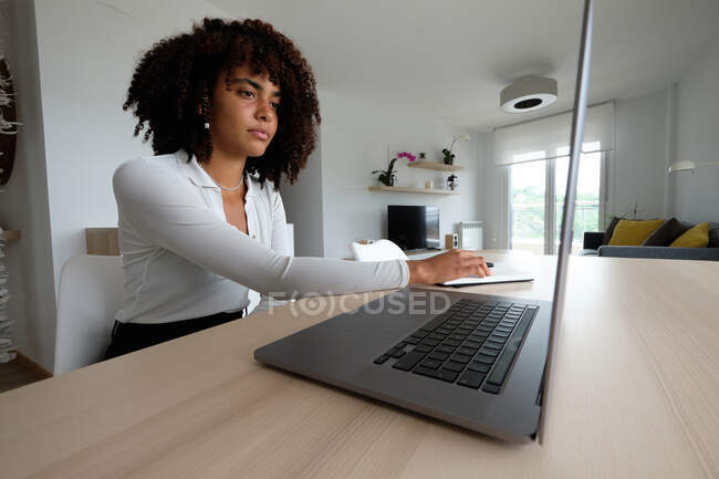 Vue latérale d'une femme afro-américaine indépendante assise à table avec un ordinateur portable et écrivant dans un bloc-notes tout en travaillant à distance sur un projet à la maison — Photo de stock