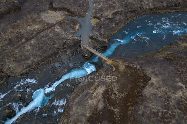 Vista desde arriba del dron del puente que cruza el arroyo rápido con agua azul en terreno accidentado en Islandia - foto de stock