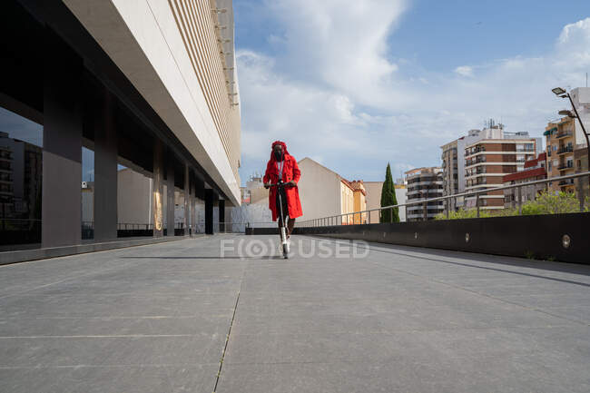 Corpo inteiro de fêmea afro-americana em máscara com tranças brilhantes montando scooter elétrico ao longo do edifício da torre no tempo livre — Fotografia de Stock