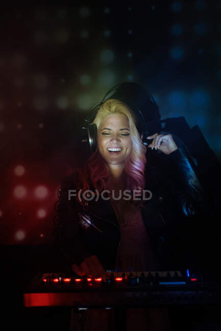 Щаслива молода дівчина-ді-джей в навушниках посміхається і використовує синтезатор для відтворення музики, стоячи в диму під час вечірки в нічному клубі — стокове фото