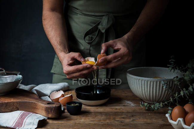 Ragazzo irriconoscibile in grembiule rompere uovo crudo sopra ciotola mentre si prepara pasticceria su tavola legname vicino stoviglie — Foto stock