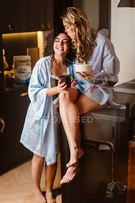 Corps complet de copine pieds nus embrassant souriant lesbienne bien-aimée tout en étant assis sur la table dans la cuisine le matin — Photo de stock