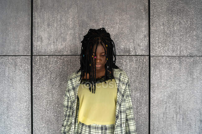 Femme afro-américaine déçue en costume à carreaux debout contre un mur gris et regardant vers le bas avec tristesse — Photo de stock