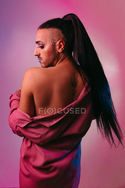 Portrait of glamorous transgender bearded woman in sophisticated make up posing against pink background at studio showing back — Stock Photo