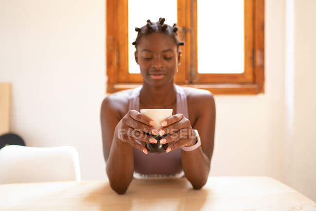 Femme afro-américaine sereine avec les yeux fermés assis à table avec une boisson rafraîchissante dans une tasse — Photo de stock