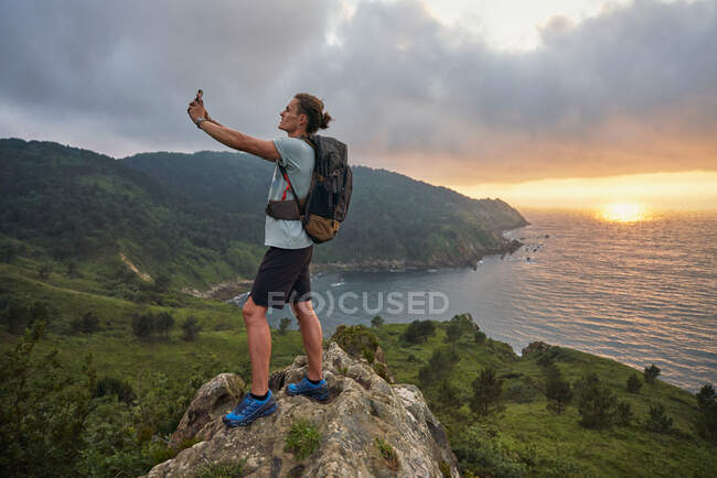 Вид сбоку на восхищенного туриста-мужчину, делающего селфи на смартфон, стоя на холме на фоне моря во время похода летом — стоковое фото