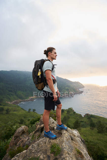 Seitenansicht eines männlichen Reisenden, der auf einem Felsen steht und beim Trekking im Sommer den Blick aufs Meer bewundert — Stockfoto