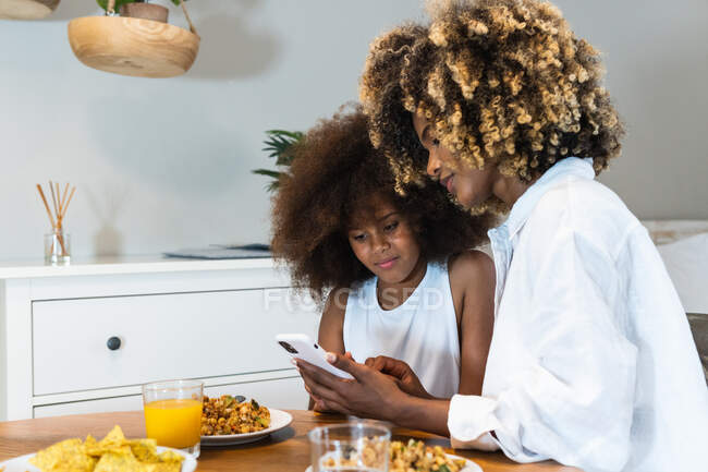 Content African American mother and teenage girl with curly hair using mobile phone together while sitting at table with food during lunch at home — Stock Photo