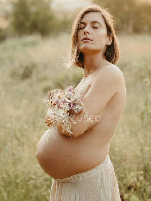 Боковой вид спокойной беременной женщины, обнаженной грудью с букетом цветов, стоящей летом в сельской местности — стоковое фото