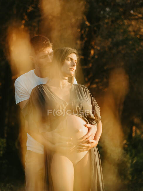 Side view of smiling male hugging pregnant female from behind while standing in countryside meadow at sunset — Stock Photo