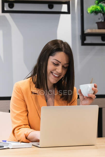 Delighted female freelancer drinking coffee and using netbook during remote work on startup in cafe — Stock Photo