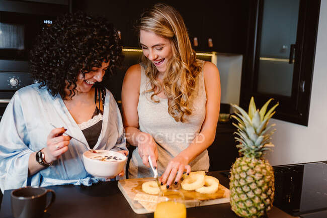 Alto ângulo de alegre casal lésbico rindo alegremente ao cortar abacaxi e comer cereais com bagas e iogurte — Fotografia de Stock