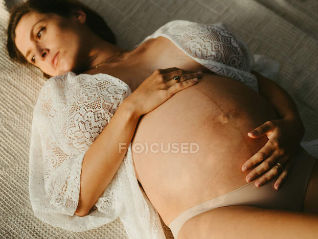Dall'alto di femmina incinta pacifica in abito trasparente toccando pancia mentre sdraiato sul letto e guardando altrove — Foto stock