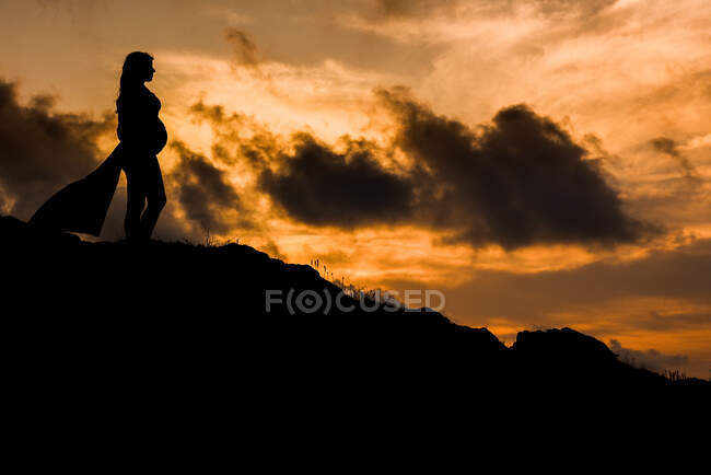 Baixo ângulo silhueta vista lateral de fêmea grávida anônima em pé sozinho na colina e sonhando com o futuro ao pôr do sol — Fotografia de Stock