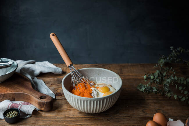 Whisk utensil in a bowl with pumpkin puree, eggs and flour for pie preparation on timber table near cutting board and towel — Stock Photo