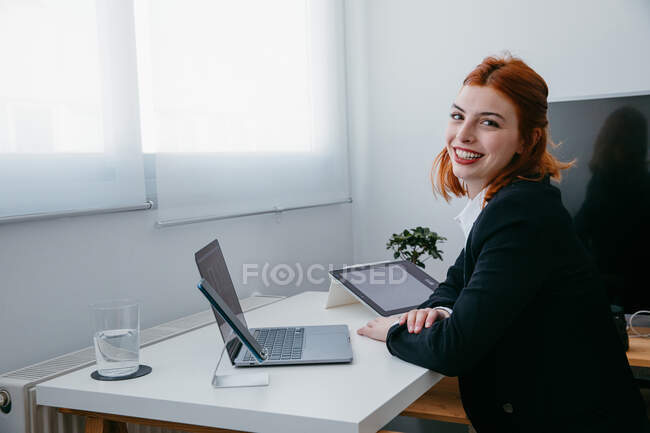 Vista lateral da jovem empresária trabalhando no netbook na mesa com tablet e smartphone em casa — Fotografia de Stock