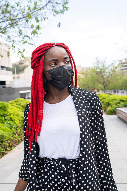 Jeune femme afro-américaine en masque de protection avec dreadlocks lumineux flânant dans le parc avec valise et détournant les yeux — Photo de stock