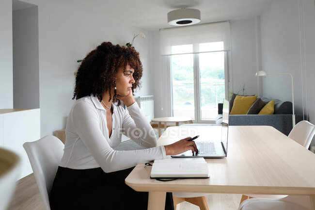 Side view of African American female freelancer sitting at table with laptop and writing in notepad while working remotely on project at home — Stock Photo