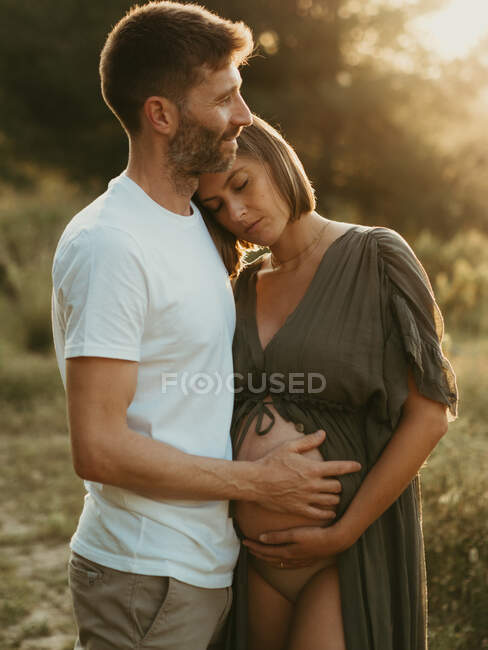 Delighted male hugging pregnant female standing in countryside at sunset enjoying summer day — Stock Photo