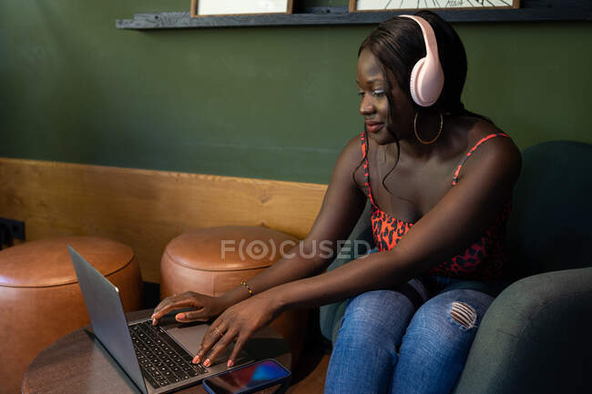 Freelancer afro-americano sorridente em fones de ouvido navegando netbook enquanto sentado no café e trabalhando no projeto — Fotografia de Stock