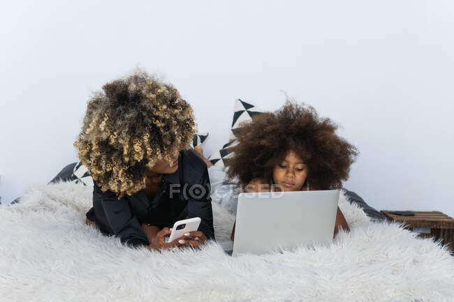 Black woman and girl browsing smartphone and laptop while resting on bed on weekend day at home together — Stock Photo