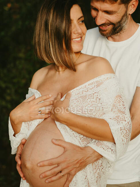 Усміхнений чоловік обіймає вагітної жінки ззаду, стоячи в сільській місцевості на заході сонця — стокове фото