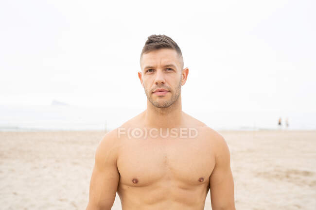 Determined male athlete with muscular naked torso standing on seashore in summer and looking forward — Stock Photo