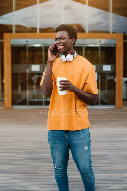 Sorridente maschio afroamericano con una tazza di caffè che ride felicemente mentre parla sul cellulare e distoglie lo sguardo — Foto stock