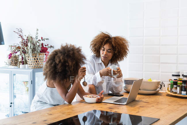 Афроамериканка и девушка просматривают социальные сети в нетбуке, сидя за кухонным столом и завтракая вместе — стоковое фото