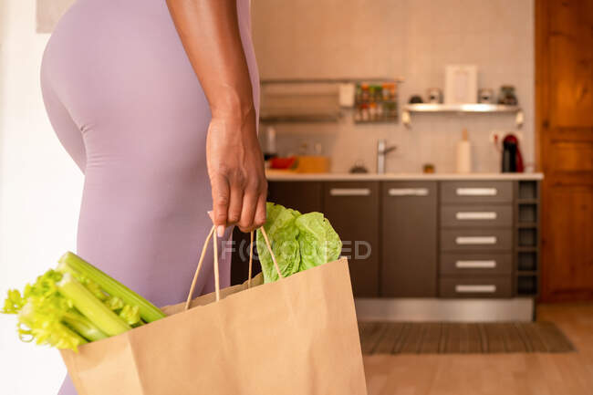 Vista lateral de la cosecha anónima hembra étnica con apio verde maduro y lechuga en bolsa de compras de pie en la cocina en casa - foto de stock