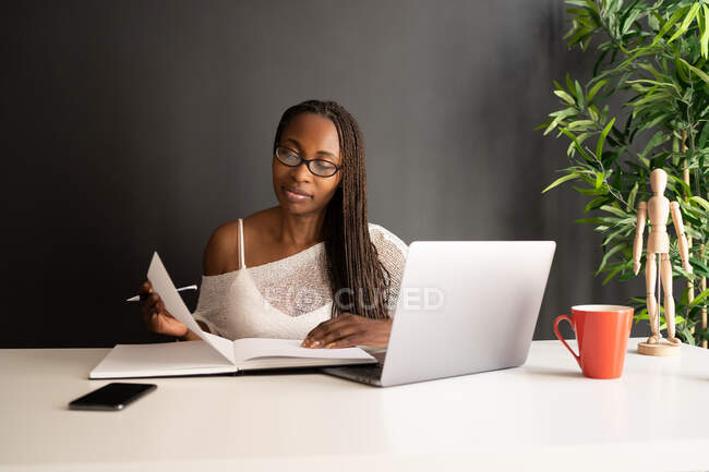 Зміст чорний фрілансер читає нотатки в блокноті, сидячи за столом з ноутбуком і працюючи над бізнес-проектом в сучасному домашньому офісі — стокове фото