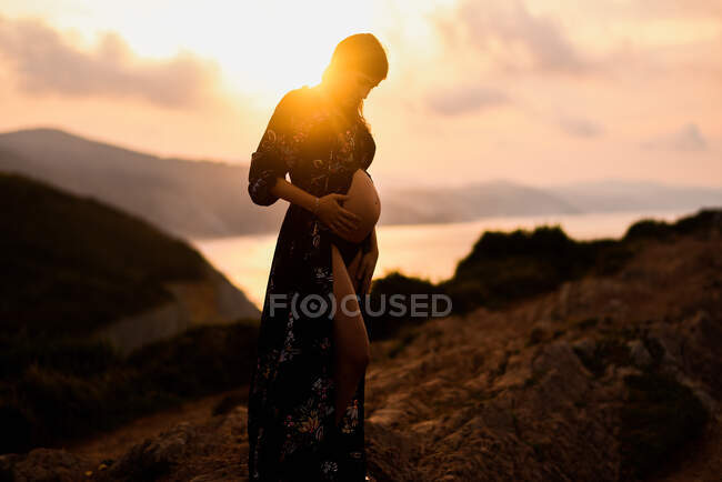 Vista lateral de la hembra embarazada parada sola en la colina y soñando con el futuro al atardecer - foto de stock