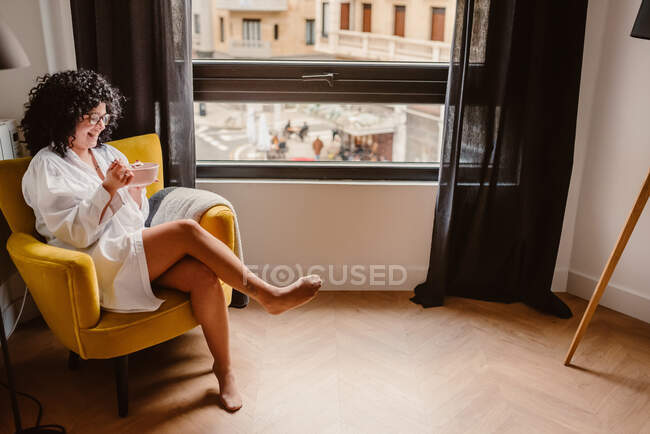 Alto angolo corpo pieno di femmina positiva negli occhiali seduto con le gambe incrociate e mangiare cibo dalla ciotola al mattino — Foto stock