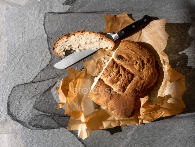 Vista superior de delicioso pão de massa de pão cozido na hora com pedaço fatiado e faca colocada no papel manteiga no fundo cinza — Fotografia de Stock