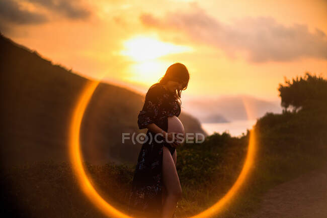 Visão lateral de amar a jovem grávida tocando barriga e sorrindo enquanto está em pé na costa montanhosa ao pôr do sol — Fotografia de Stock